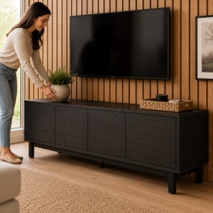Peut inclure: Un meuble TV en bois brun foncé avec quatre portes et des pieds noirs. Une femme place une plante en pot sur le dessus. Une grande télévision noire est fixée au mur en bois derrière. Un plateau tressé est posé sur le meuble.