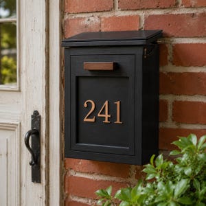 May include: A black mailbox with a wooden handle and the number 241 in copper numerals. The mailbox is mounted on a red brick wall. The top of the mailbox is slightly open.