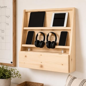 May include: A light-colored wooden charging station with multiple shelves. It holds two tablets, three smartphones, and two pairs of black headphones. The station is mounted on a white wall, with a calendar visible on the left.