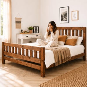 May include: A wooden bed frame with a headboard and footboard. The bed is made of stained wood and has a simple, rustic design. The bed is covered with a white comforter, brown throw blanket, and decorative pillows. A person is sitting on the bed.