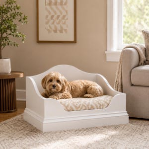 May include: A white dog bed with a curved headboard and footboard. The bed has a patterned cushion and a small dog lying inside. The dog has light brown fur and is looking at the camera.