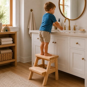 May include: A two-step wooden stool in a bathroom setting. The stool is made of light-colored wood and is designed to help a child reach the sink. The child is standing on the stool, washing their hands at a gold faucet.