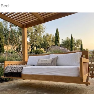 May include: A wooden swing bed with white bedding and pillows hangs from a wooden pergola. A decorative throw blanket and a pillow with the word "love" are on the bed. The bed is suspended by rope.