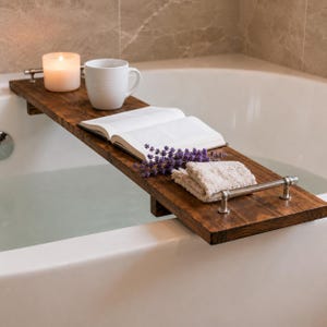 May include: A dark stained wooden bathtub tray with a lit candle, white mug, open book, lavender sprigs, and a folded towel. The tray has a metal handle and rests across a white bathtub filled with water.
