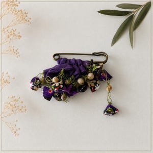 May include: A bronze-toned safety pin brooch adorned with gathered purple fabric, floral patterned fabric scraps, small wooden beads, and thin green wire accents. The decorative cluster hangs from the metal pin, featuring a mix of solid and printed textiles.