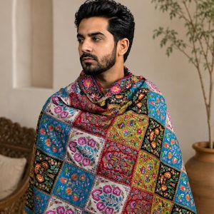Puede incluir: Un chal rectangular con un vibrante diseño de patchwork de paneles cuadrados. Cada cuadrado muestra un intrincado bordado floral en tonos azul, rojo, amarillo y negro. La tela está drapeada sobre los hombros, mostrando un patrón repetitivo de motivos botánicos y flores coloridas en toda la superficie.