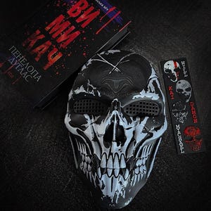 May include: A black and white skull mask with mesh eye coverings, positioned next to a book titled "ВИ МИ КАУ" by Penelope Douglas and a rectangular bookmark featuring four distinct skull illustrations labeled Will, Kai, Damon, and Michael.