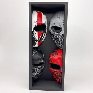 May include: A rectangular black display box containing four half-face skull masks. The masks feature distinct color schemes: one white with a red stripe, one silver with black weathering, one matte black with grey details, and one bright red with black accents.