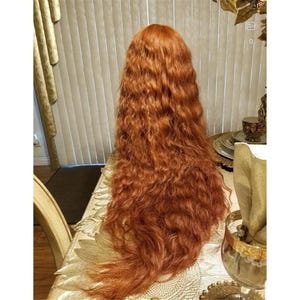 May include: An auburn wig with long, wavy hair. The wig is displayed on a table, with the hair flowing down. The colour is a rich red-orange, and the curls are clearly defined. The wig's length is considerable.