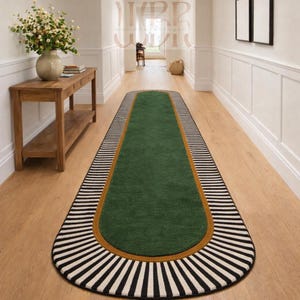 May include: A green runner rug with a black and white striped border. The rug has a gold-colored trim and a gray border. The rug is placed on a wooden floor in a hallway.