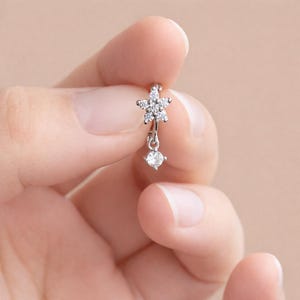 May include: A silver earring featuring a star-shaped design with five clear, sparkling stones. Below the star hangs a single, round, clear stone. The earring is held between fingers against a soft, neutral background.