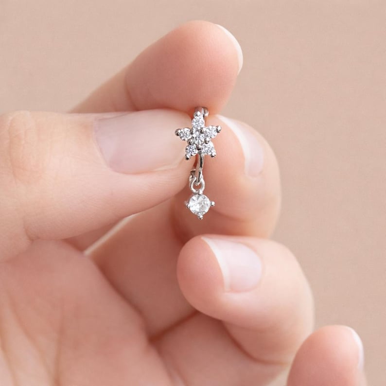 May include: A silver earring featuring a star-shaped design with five clear, sparkling stones. Below the star hangs a single, round, clear stone. The earring is held between fingers against a soft, neutral background.