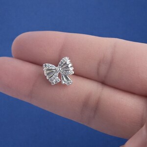 May include: A small, silver-colored bow-shaped charm with clear, sparkling stones. The charm is highly detailed and appears to be made of metal. The bow is centered in the image against a blue background.