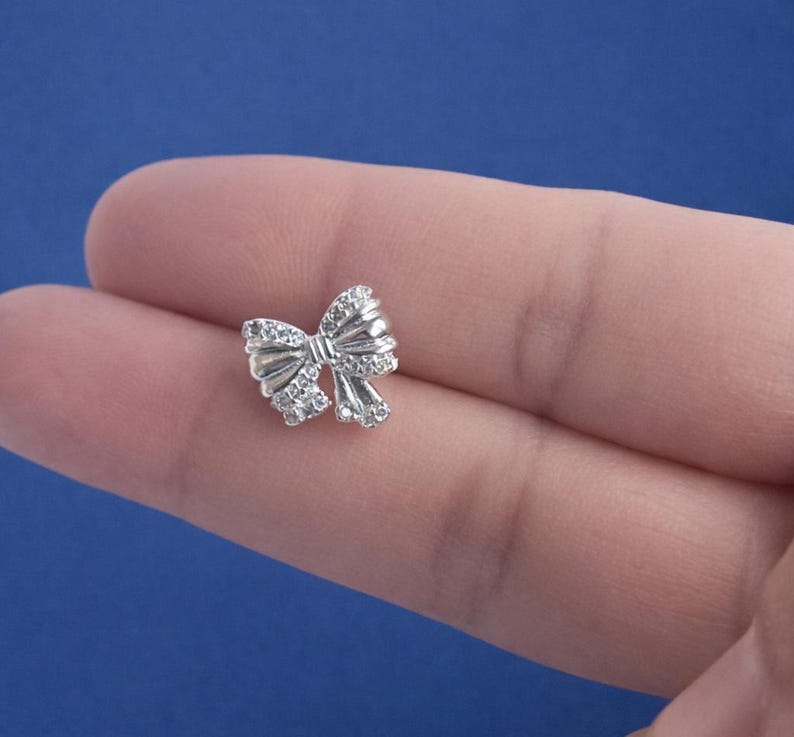 May include: A small, silver-colored bow-shaped charm with clear, sparkling stones. The charm is highly detailed and appears to be made of metal. The bow is centered in the image against a blue background.