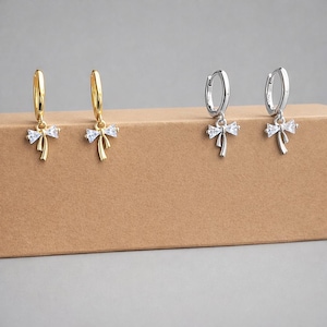 May include: Four earrings are shown, two gold and two silver. Each has a bow pendant with a clear stone. The earrings are on a brown surface, against a neutral background. The bow design is a delicate detail.