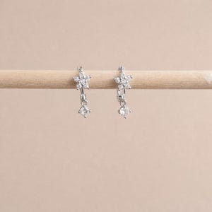 May include: A pair of silver-toned earrings. Each earring features a flower-shaped top with multiple small clear stones. A single clear, round stone dangles below each flower. The earrings are displayed on a light-colored wooden dowel against a neutral background.