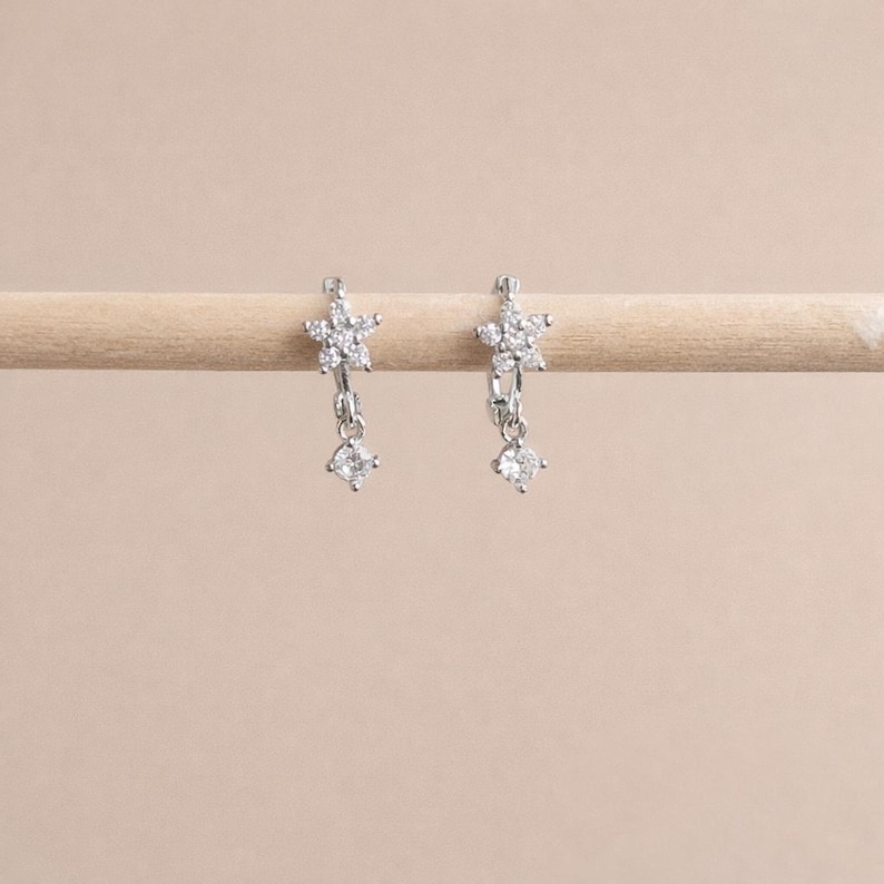 May include: A pair of silver-toned earrings. Each earring features a flower-shaped top with multiple small clear stones. A single clear, round stone dangles below each flower. The earrings are displayed on a light-colored wooden dowel against a neutral background.