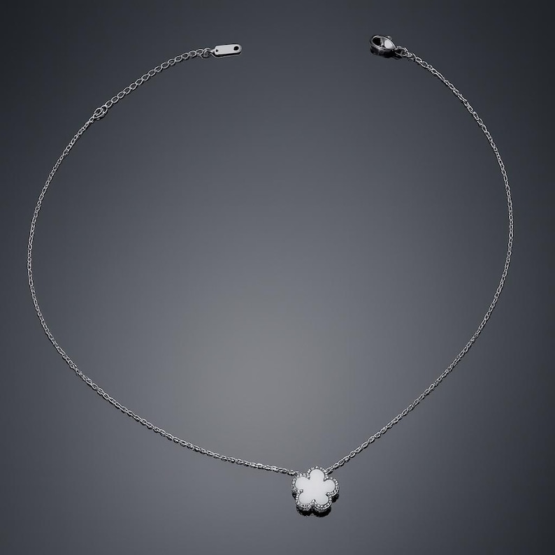 Op de afbeelding: Een delicate zilveren ketting met een bloemvormige hanger. De hanger heeft een wit midden en is omlijnd met kleine, sprankelende accenten. De ketting is dun en elegant, met een eenvoudige sluiting.