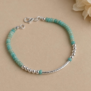 May include: A delicate bracelet with small, round, turquoise beads and silver beads, plus a textured silver bar. The bracelet fastens with a silver clasp. The jewellery is displayed on a neutral background.