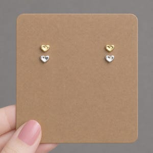 Op de afbeelding: Twee paar hartvormige oorstekers, één goudkleurig en één zilverkleurig, gepresenteerd op een bruine kaart. De oorbellen zijn klein en minimalistisch. De kaart wordt door een persoon vastgehouden.