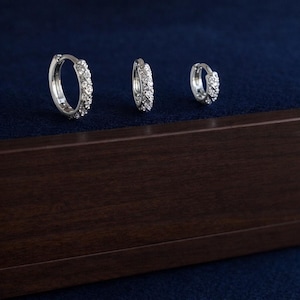 May include: Three silver hoop earrings, each adorned with a row of small, clear gemstones. They are presented on a dark blue surface, with a dark brown wooden base. The earrings are of varying sizes.