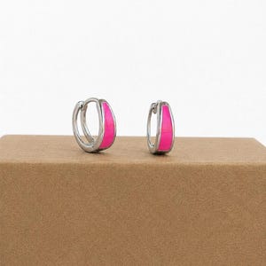 May include: A pair of small silver hoop earrings featuring a vibrant hot pink enamel inlay on the front surface. The earrings have a hinged closure mechanism and a polished metallic finish, presenting a sleek and compact design for everyday wear.