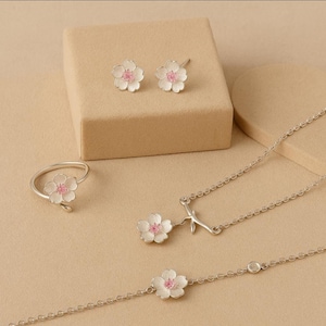 Peut inclure: Un ensemble de bijoux comprenant une bague, des boucles d'oreilles, un collier et un bracelet. Chaque pièce est en argent et présente un motif de fleur blanche avec un centre rose. Les boucles d'oreilles sont des clous et le bracelet a un petit accent rond.