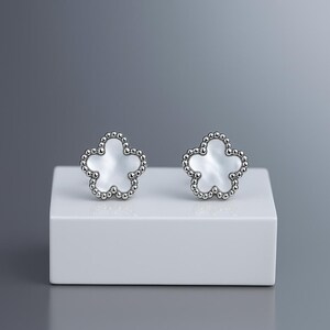 Op de afbeelding: Een paar zilverkleurige bloemvormige oorstekers. Elke oorbel heeft een witte, parelmoerachtige kern en een zilveren kralenrand. De oorbellen worden gepresenteerd op een wit, rechthoekig platform tegen een grijze achtergrond.