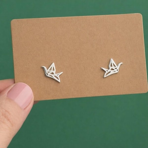 May include: Silver origami bird stud earrings on a brown card. The earrings feature a geometric bird design. The background is a solid green colour.