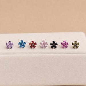 May include: Seven flower-shaped stud earrings arranged in a row, each featuring five pear-cut gemstones in purple, blue, red, light pink, black, dark pink, and olive green, all set in a silver-toned metal frame with a central circular stone.