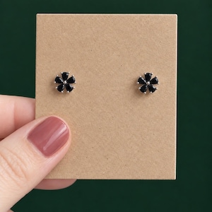 May include: A pair of stud earrings featuring five black teardrop-shaped stones arranged in a flower pattern. The stones are set in silver-toned metal prongs and are mounted on a square piece of light brown cardboard for display purposes.