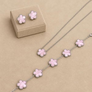 Può includere: Un set di gioielli con collana, bracciale e orecchini a lobo. Ogni pezzo ha una montatura in metallo color argento con un centro rosa a forma di fiore. Gli orecchini sono su un portagioie beige.