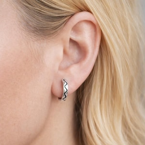 Peut inclure: Une boucle d'oreille argentée avec un motif ondulé noir. La boucle d'oreille est portée sur une oreille, mettant en valeur les détails et le design du bijou. La boucle d'oreille est de forme circulaire.