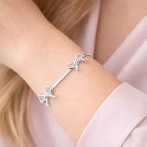 Puede incluir: Una pulsera plateada con dos dijes en forma de lazo. La pulsera tiene un diseño delicado, con los lazos colocados a ambos lados de la muñeca. La pulsera se muestra en el brazo de una persona, sobre un fondo rosa suave.