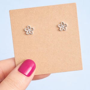 May include: A pair of silver flower stud earrings on a tan card. The earrings have a delicate, openwork design. The background is light blue. The earrings are small, suitable for everyday wear.