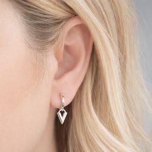 以下が含まれることがあります： シルバーのフープピアス。幾何学的なV字型のペンダントが付いています。 金髪の背景に耳に装着されたピアスが写っています。 シンプルでモダンなデザインで、普段使いに最適です。