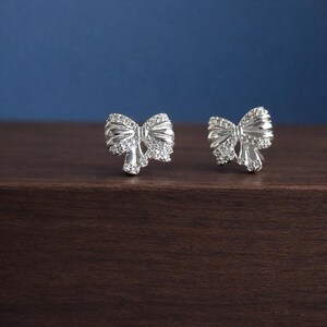 May include: A pair of silver bow-shaped stud earrings. Each earring features a detailed bow design, embellished with small, sparkling stones. The earrings are displayed on a dark wooden surface against a blue background, highlighting their elegant design.