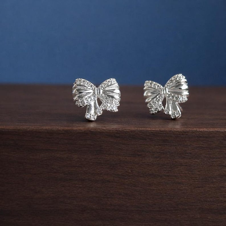 May include: A pair of silver bow-shaped stud earrings. Each earring features a detailed bow design, embellished with small, sparkling stones. The earrings are displayed on a dark wooden surface against a blue background, highlighting their elegant design.