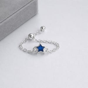 May include: A silver adjustable ring with a blue star-shaped gemstone. The ring has a delicate chain design and two small silver beads. The star is the focal point, set against a white background.