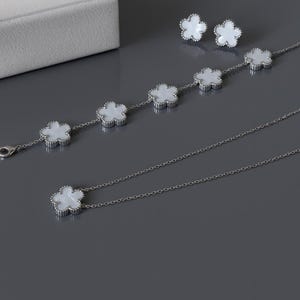 Op de afbeelding: Een sieradenset met een ketting, armband en oorbellen. Elk stuk heeft een zilveren ketting en witte bloemvormige hangers. De oorbellen zijn studs en de armband heeft vijf bloemvormige bedels. De ketting heeft &eacute;&eacute;n bloemvormige bedel.