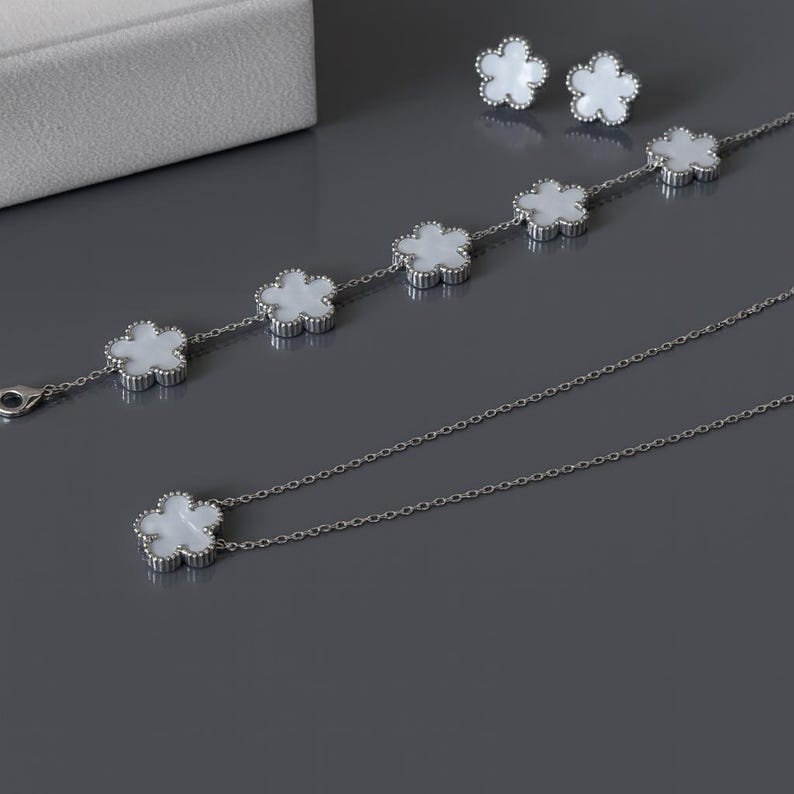 Op de afbeelding: Een sieradenset met een ketting, armband en oorbellen. Elk stuk heeft een zilveren ketting en witte bloemvormige hangers. De oorbellen zijn studs en de armband heeft vijf bloemvormige bedels. De ketting heeft &eacute;&eacute;n bloemvormige bedel.