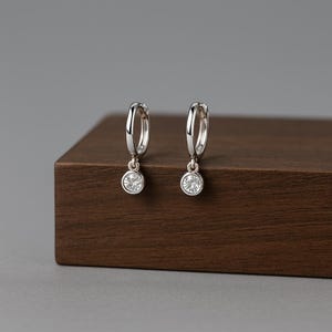 Puede incluir: Un par de pendientes de aro plateados, cada uno con una pequeña gema redonda y transparente colgando. Los pendientes se exhiben sobre una superficie de madera marrón oscuro, con un fondo gris. Diseño sencillo y elegante.