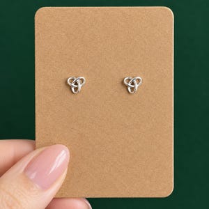 May include: A pair of silver-coloured stud earrings featuring a triquetra knot design. The earrings are mounted on a rectangular piece of light brown cardstock, which provides a neutral display for the metallic, interlocking circular shapes.