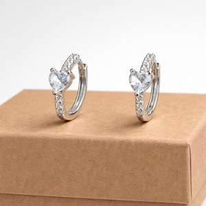 May include: A pair of silver hoop earrings. Each earring features a heart-shaped, clear gemstone and is accented with small, clear stones. The earrings are displayed on a brown surface against a white background.