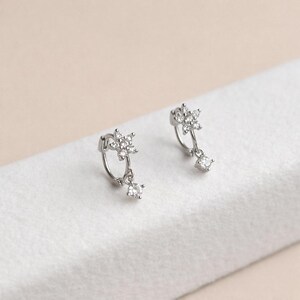 May include: A pair of silver hoop earrings. Each earring features a star-shaped cluster of small, clear crystals at the top, with a single square-cut crystal dangling below. The earrings are displayed on a white surface.