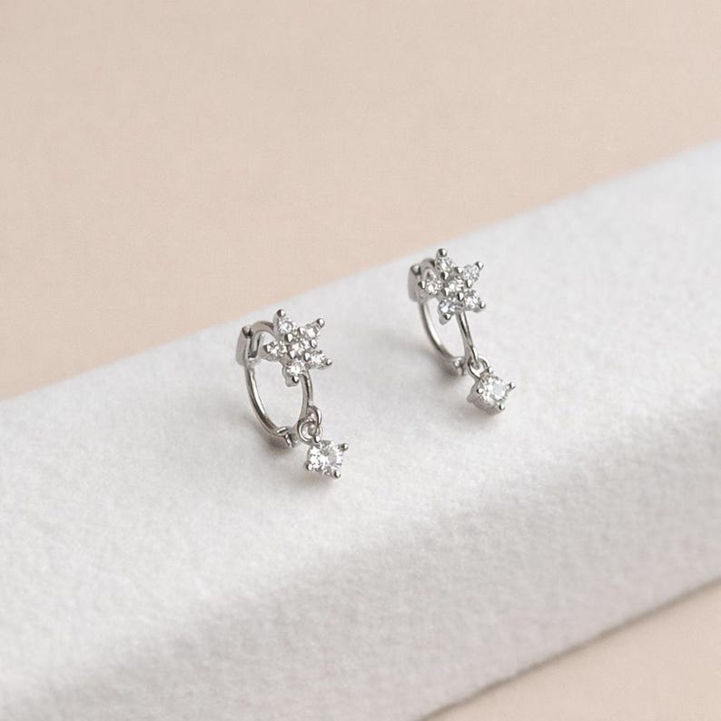 May include: A pair of silver hoop earrings. Each earring features a star-shaped cluster of small, clear crystals at the top, with a single square-cut crystal dangling below. The earrings are displayed on a white surface.