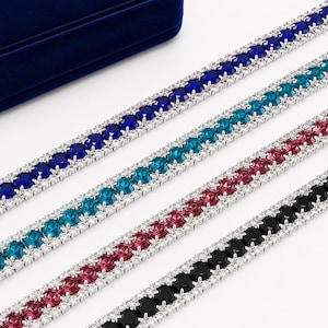 May include: Four tennis bracelets featuring a central row of round-cut stones in royal blue, turquoise, magenta, and black, each flanked by two rows of smaller clear white stones set in a metallic frame, arranged diagonally across the frame.