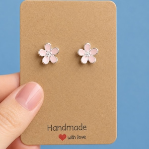 以下が含まれることがあります： ピンクの花びらと銀色の縁取りが施された、花の形をしたスタッドピアス。 「Handmade with love」と書かれた茶色のカードに展示されています。 背景は無地の青色です。