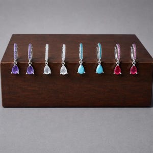 Puede incluir: Una colección de siete pendientes de aro plateados, cada uno con una piedra preciosa en forma de lágrima de diferentes colores. Las piedras preciosas incluyen púrpura, transparente, turquesa y rosa. Se exhiben sobre un bloque de madera marrón oscuro.