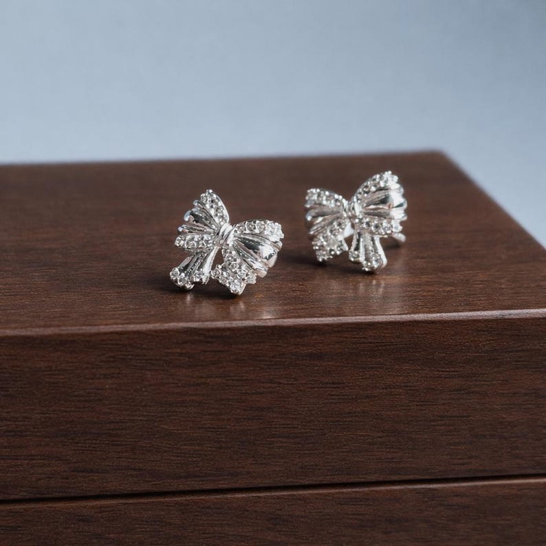May include: A pair of silver-colored bow-shaped stud earrings. Each earring is adorned with small, sparkling crystals, creating a shimmering effect. The earrings are displayed on a dark brown wooden surface, with a soft blue background.